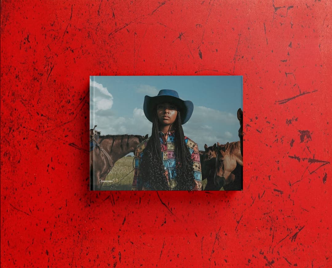 The image shows a woman standing in front of a vibrant red background with a distressed or grunge texture. She is wearing a blue cowboy hat and a colorful, patterned shirt. Behind her, a group of horses are visible, including a brown horse and a lighter brown or tan horse. The woman has long, dark hair. The image is fr