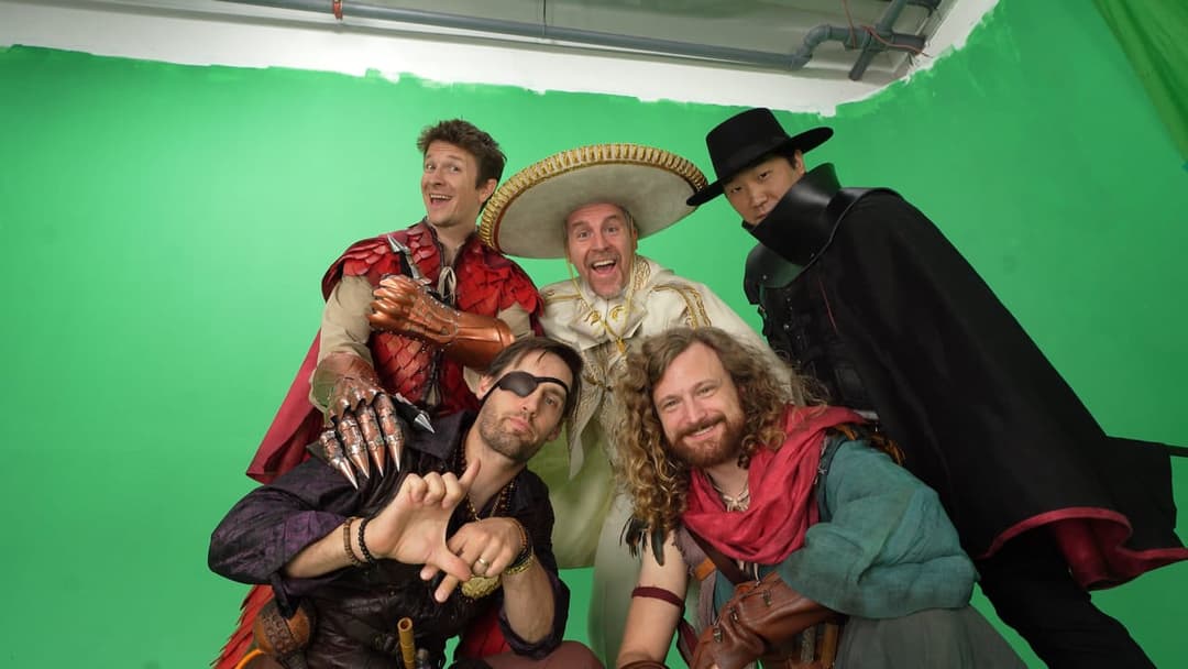 The image shows five men posing against a vibrant green backdrop, likely a green screen used in film or television production. The man on the far left is wearing a red and gold outfit, including a cape and a sombrero. Next to him, a man in a white outfit with a sombrero is smiling. The third man from the left is wearin