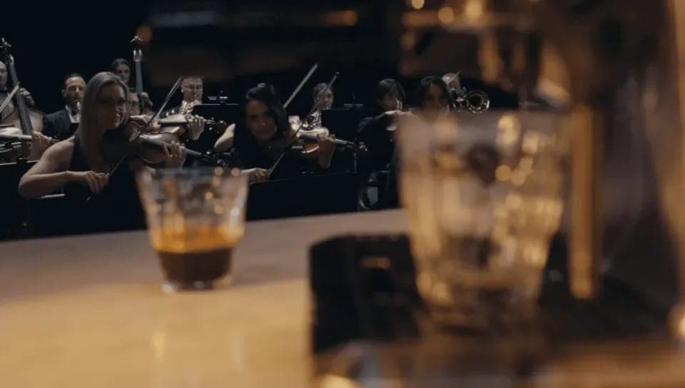 The image shows a group of musicians, likely a string quartet, performing on a stage. The musicians are seated in a row, with their instruments held in their hands. The stage is dark, and the musicians are out of focus, making it difficult to discern the exact instruments they are playing. In the foreground, a glass of