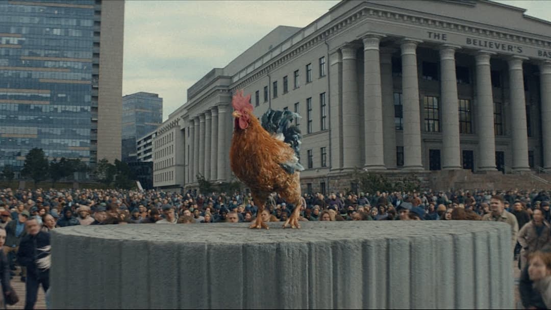 A vibrant orange and white rooster stands proudly on a large, gray stone pedestal in the center of a bustling city square. The rooster's head is turned to the left, and its tail feathers are spread out. The pedestal is surrounded by a crowd of people, many of whom are looking towards the rooster. In the background, a l