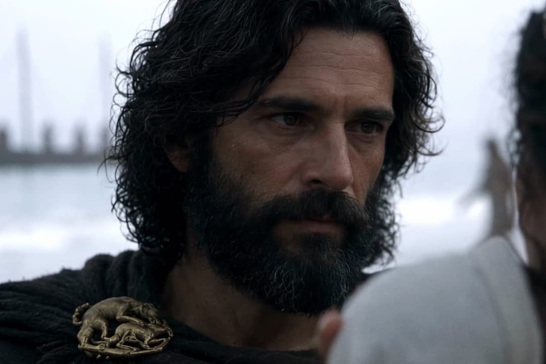 The image shows a man with long, dark hair and a beard, wearing a dark-colored garment with a gold-colored brooch featuring a stylized animal design. He is looking off to the side, and his expression is serious. The background is out of focus, but appears to be a body of water with a hazy or foggy atmosphere. The man's