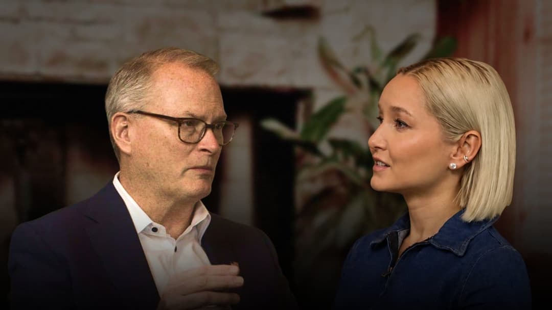 The image shows a man and a woman engaged in a conversation. The man, wearing a dark suit and glasses, is on the left side of the frame. The woman, with blonde hair, is on the right side of the frame. They are standing in front of a brick wall with a plant in the background. The man is gesturing with his hands, while t