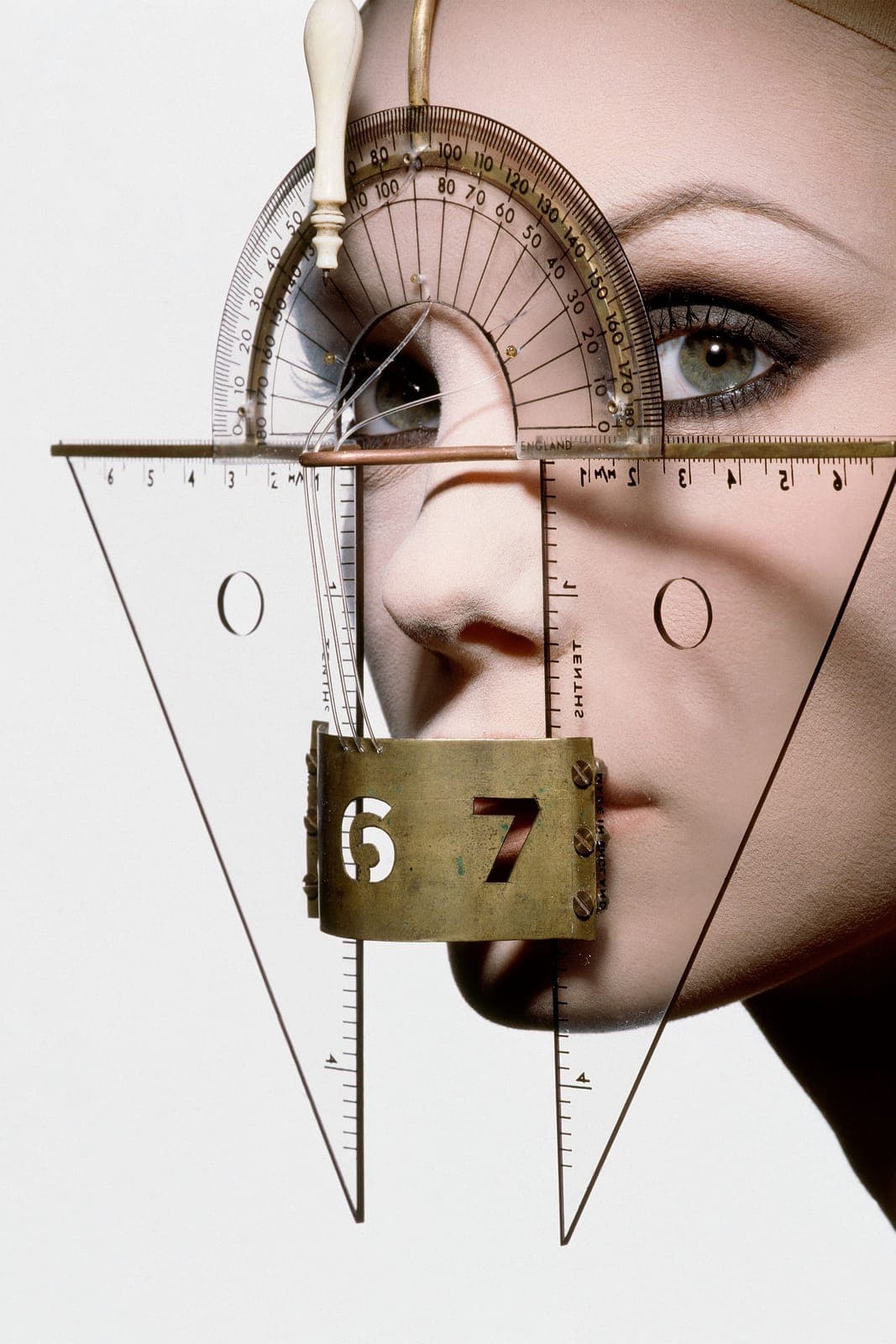 The image shows a close-up of a mannequin head with a unique design. The mannequin's face is adorned with a gold-colored metal frame that holds a series of metal rods and a metal ruler. The ruler is marked with numbers from 0 to 180, and the mannequin's eyes are visible through a small hole in the frame. The mannequin'