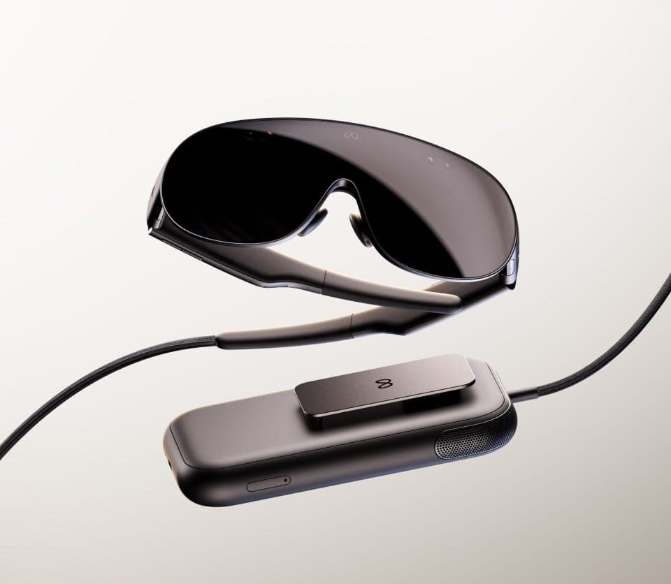 The image presents a pair of sleek, black-framed glasses suspended in mid-air, with the lenses facing upwards. The glasses are connected to a black rectangular device, which appears to be a charging case, by a black cord. The charging case has a small, dark-colored button on its side. The background is a light beige or