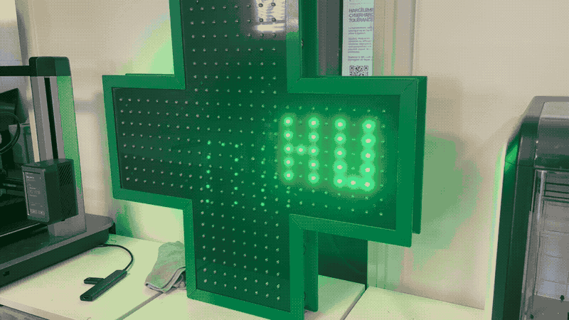 The image shows a green cross-shaped sign with a grid of small, round, light-colored dots, possibly LEDs, on its surface. The sign is illuminated with a bright green light, and the letters "HU" are prominently displayed in the center. The sign is positioned on a white surface, possibly a table or counter, and is surrou