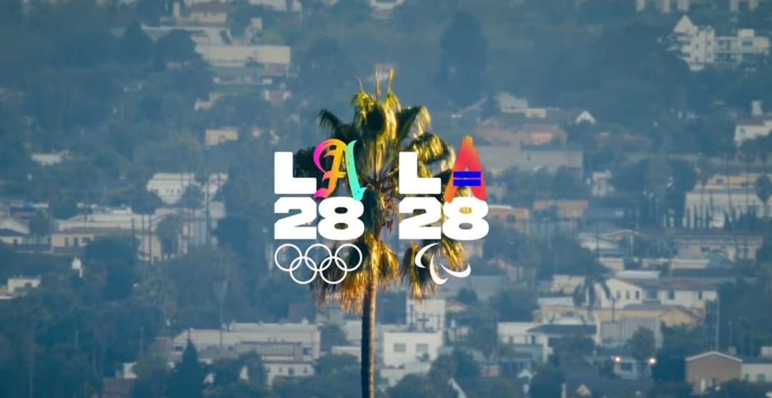 A blurred hazy city view with a tall palm tree in the center, overlaid with the LA28 logo in white and multicolor script, the Olympic rings below the left "28" and the Paralympic symbol below the right "28".