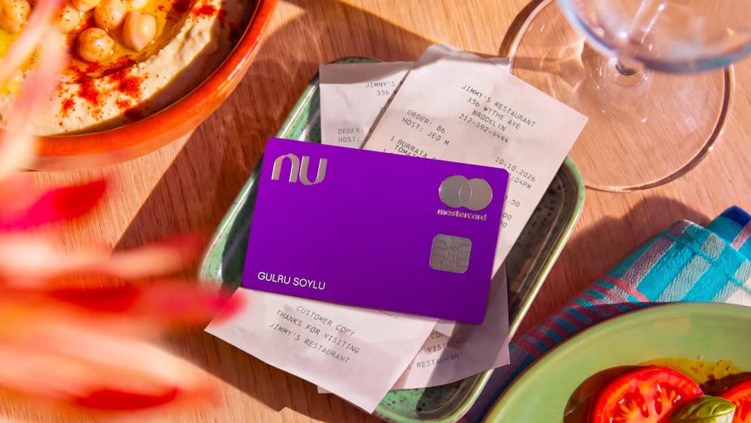 A purple Nu credit card with Mastercard branding and the name 'GULRU SOYLU' rests on a green tray with receipts from 'Jimmy's Restaurant', next to a hummus bowl, a tomato plate, and a wine glass on a wooden table.