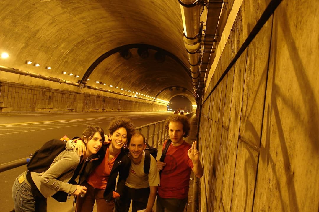 The image shows four individuals standing in a tunnel, with a concrete wall on the right and a concrete ceiling on the left. The tunnel is illuminated by a series of lights along the ceiling, casting a warm glow on the scene. The individuals are positioned in a loose line, with the person on the far left wearing a gray