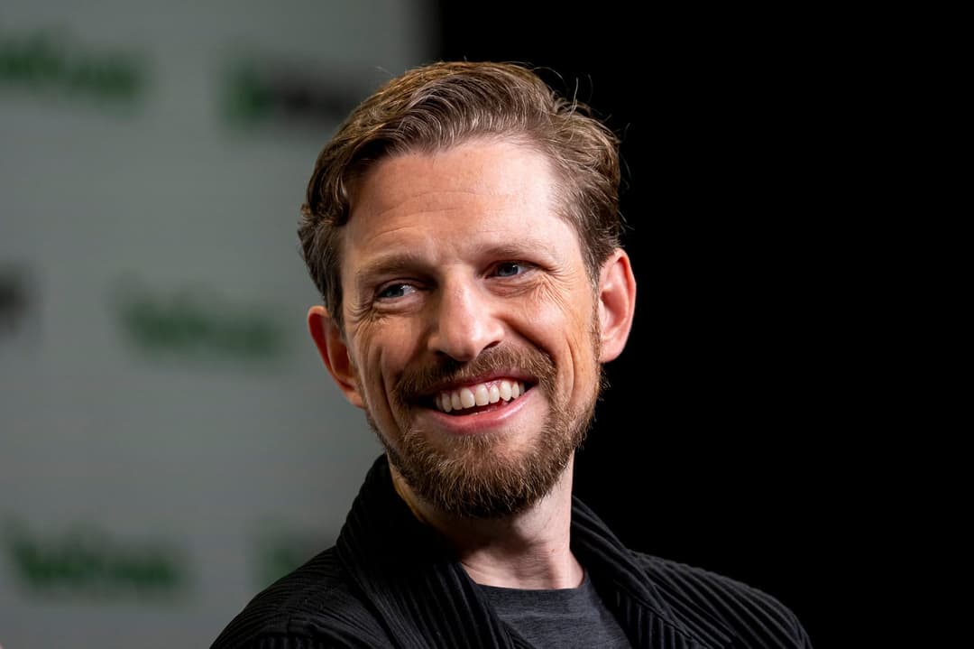 The image shows a man with light brown or blond hair and a beard, wearing a black jacket over a dark gray or black t-shirt. He is smiling and looking off to the side, with his head slightly tilted. The background is a dark, out-of-focus color, possibly black, with a blurred green and white pattern.