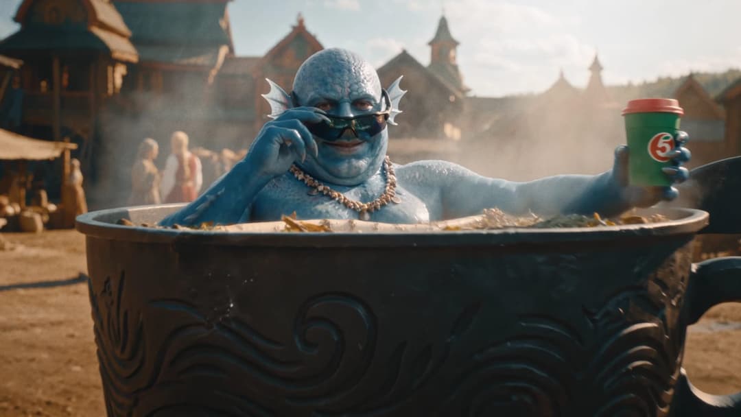 In the image, a man with blue skin and fins is seated in a large, dark-colored, possibly ceramic, bowl or pot. He is holding a green coffee cup with a red lid in his right hand and wearing sunglasses. The man is also wearing a necklace made of seashells. The bowl/pot is situated in a setting that appears to be a villag