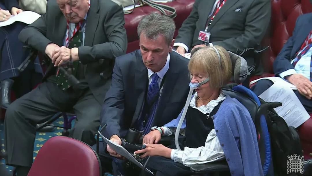 The image shows a man in a suit and tie seated in a wheelchair, with a woman in a wheelchair beside him. The man is holding a clipboard and appears to be reading or reviewing something on it. The woman is wearing a light blue cardigan over a white blouse and has a medical device connected to her face. The setting appea