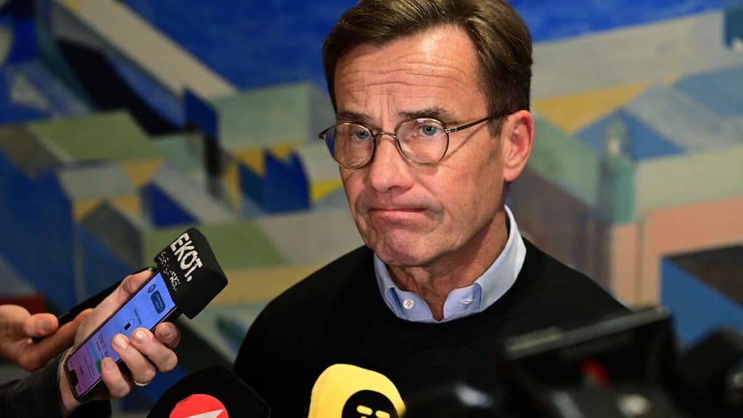 The image shows a man with short, dark brown hair and glasses, wearing a black sweater over a light blue collared shirt. He is holding a black microphone with a white logo that reads "EKOT" and a smartphone in his right hand. The smartphone displays a purple and white interface. The man is standing in front of a vibran