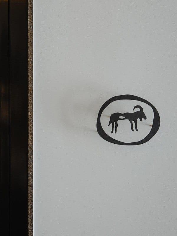The image shows a white wall with a black, hand-drawn, oval-shaped logo featuring a black silhouette of a ram or goat. The logo is positioned in the top right corner of the image. The wall is framed by a dark brown or black border, which appears to be made of wood or a similar material.
