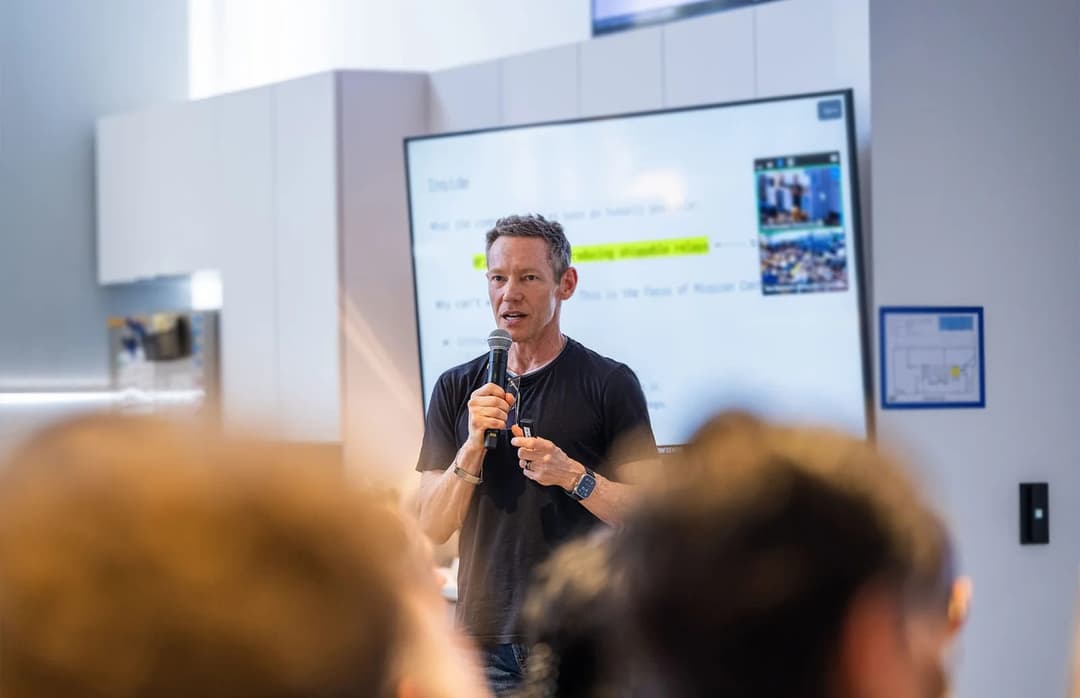 The image shows a man standing in front of a large whiteboard, addressing a group of people in a room with white walls and a light gray/beige ceiling. The man is dressed in a black t-shirt and is holding a microphone, suggesting he is giving a presentation or speech. The whiteboard displays text and a picture, possibly
