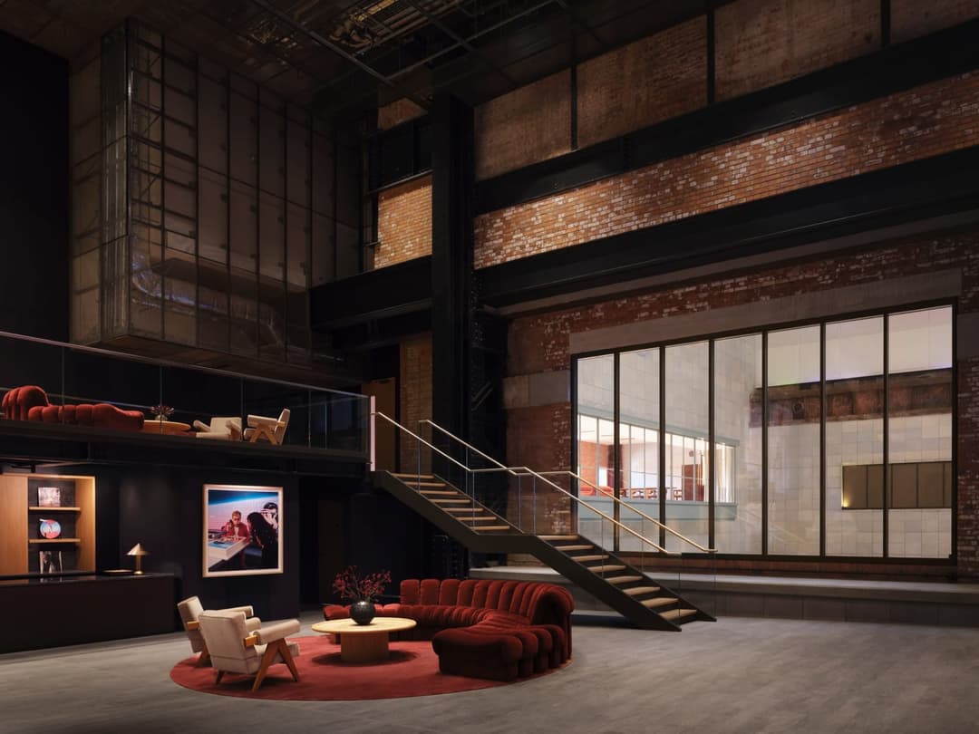 The image depicts a modern interior space with a high ceiling and exposed brick walls, creating a rustic aesthetic. A black metal staircase with a white railing ascends to a second level, which features a black railing and a dark-colored counter or bar area. A red sofa and two white armchairs are arranged on a red rug