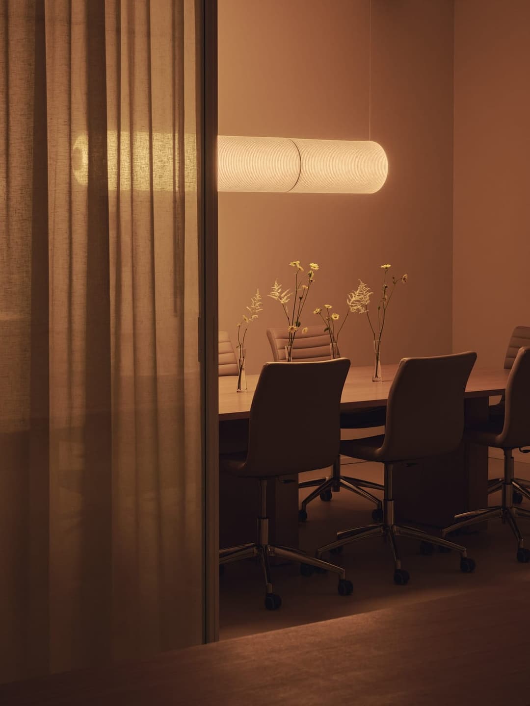 The image depicts a modern conference room with a long table draped in a white tablecloth. The table is surrounded by six chairs, each with a sleek black armrest and a light-colored, possibly beige or cream, seat. A white, cylindrical pendant light hangs above the table, casting a warm glow on the scene. The walls are