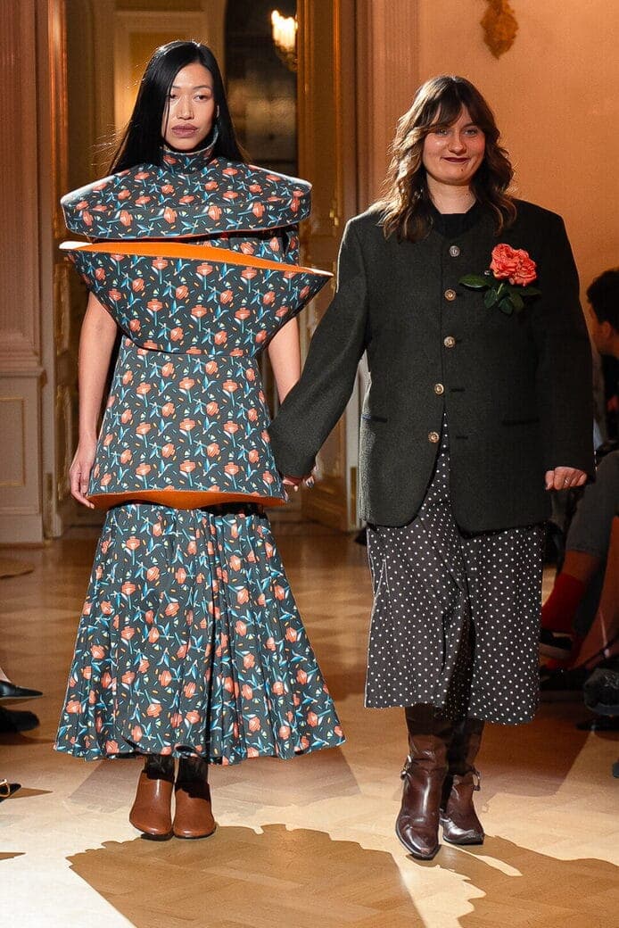 The image shows two women walking down a wooden runway. The woman on the left is wearing a vibrant dress with a floral pattern in shades of blue and orange, and a large orange circular piece attached to the dress. The woman on the right is wearing a dark green or black blazer over a black and white polka dot dress and