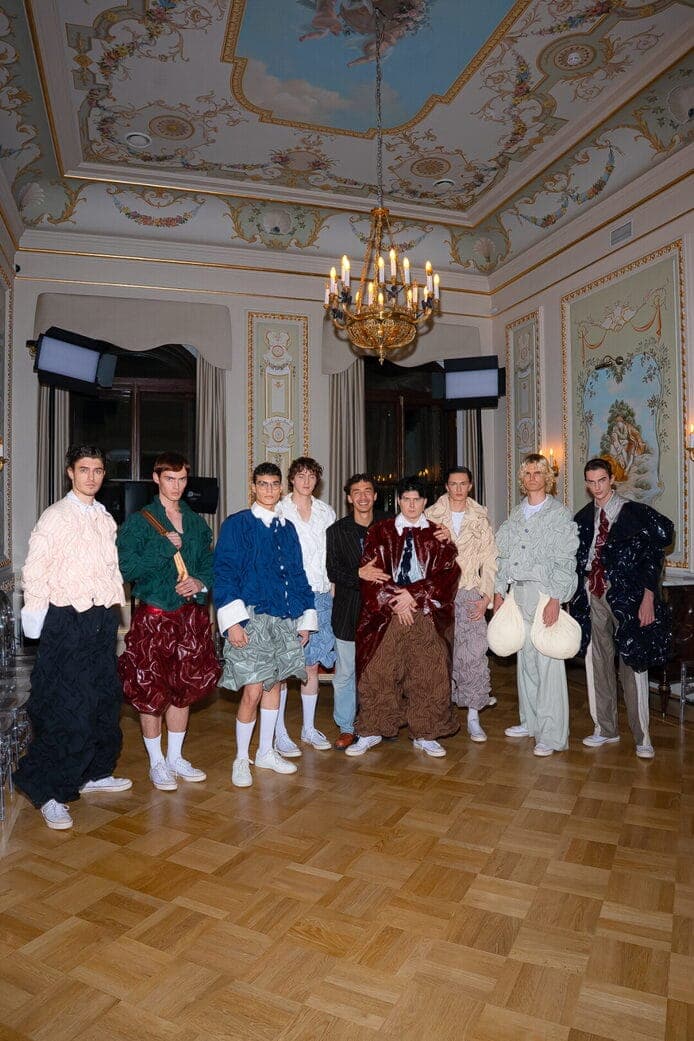 The image shows a group of nine individuals standing in a room with ornate, light-colored walls and a wooden parquet floor. The room features a large, ornate chandelier hanging from the ceiling, which is decorated with intricate patterns and designs. The walls are adorned with paintings and other decorative elements. T