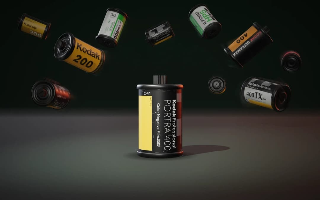 The image presents a collection of Kodak film cartridges, each with a unique color and size, scattered across a dark gray/black surface. The cartridges are arranged in a loose circular pattern, with some overlapping each other. The largest and most prominent cartridge is centrally located, featuring a black body with a