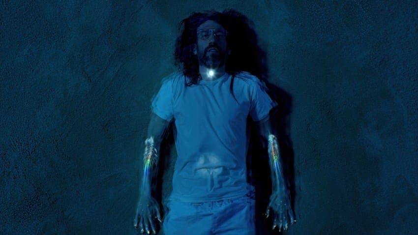 The image shows a man lying on his back on a dark blue wall. He is wearing a blue t-shirt and blue shorts. His arms are raised, and his hands are slightly spread apart. The man has long, dark hair and a beard. The image is taken from a low angle, making the man appear larger than life. The man's arms are adorned with c