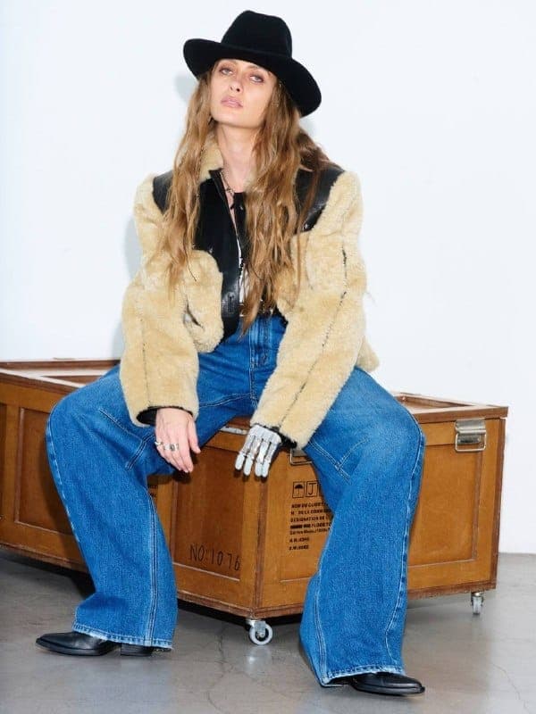 The image shows a young woman seated on a wooden crate. She is wearing a black cowboy hat, a beige fur coat, a black leather-like jacket, and blue jeans. She is also wearing black cowboy boots. The crate is positioned against a white wall, and the woman is looking off to the side. The crate has a label with the number
