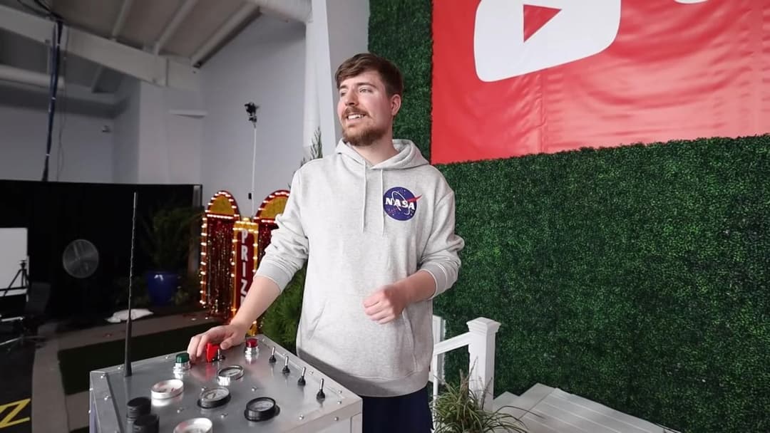 The image shows a man standing in front of a vibrant red and white YouTube logo banner. He is wearing a gray sweatshirt with a NASA logo on the left chest and dark-colored pants. He is holding a control panel with various knobs and buttons, suggesting he is operating a device. The control panel is situated on a white p