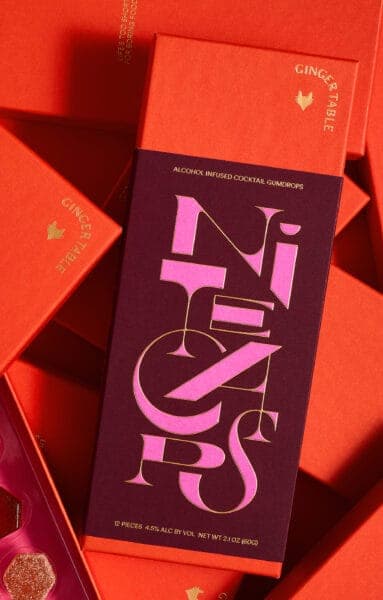 The image shows a collection of orange boxes, each adorned with a pink and gold logo that reads "GINGER TABLE" and "ALCOHOL INFUSED COCKTAIL GUMDROPS". The boxes are stacked on top of each other, creating a sense of depth and perspective. The boxes are arranged in a way that suggests they are part of a larger set, with