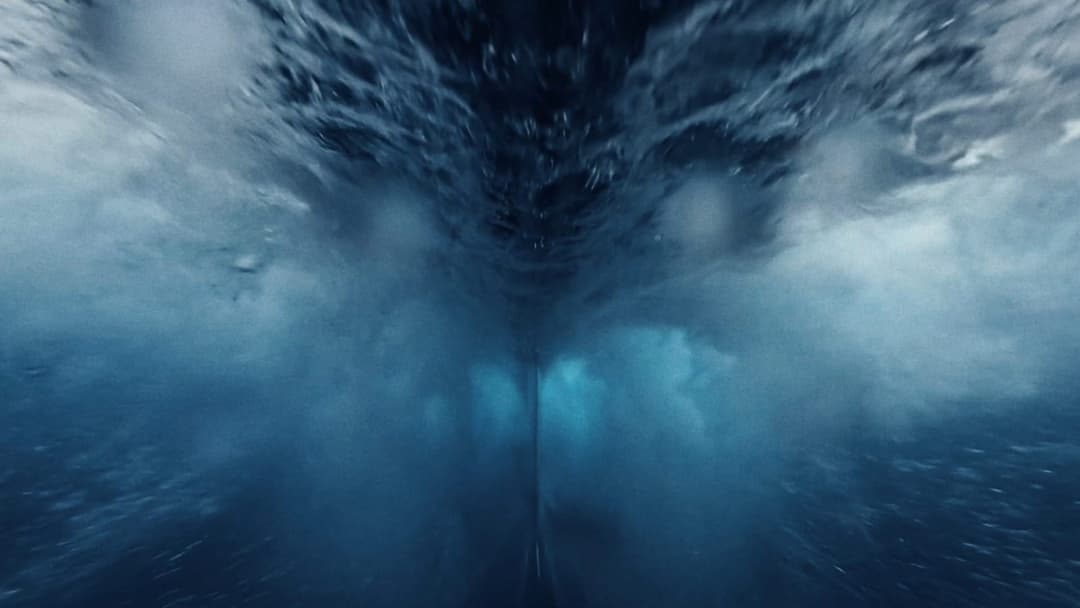 The image captures a view from the ocean floor, looking upwards towards the surface. The water is a deep blue color, with a lighter blue hue near the surface. The surface is disturbed by a large wave, creating a sense of motion and energy. The perspective is from a low angle, emphasizing the height of the wave and the