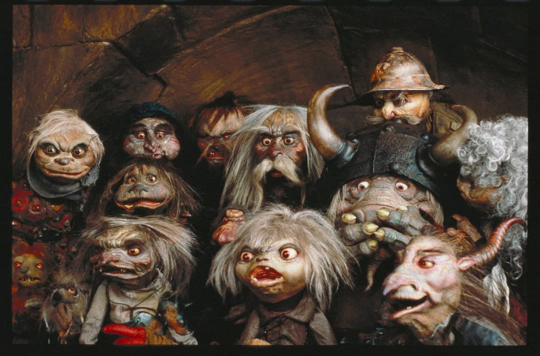 The image depicts a group of puppets, likely from a film or theatrical production, arranged in a loose grouping against a dark, textured background that resembles a stone or brick wall. The puppets are primarily white with gray hair and some have red or brown accents. They are dressed in a variety of clothing, includin