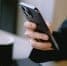 The image shows a person's hand holding a black smartphone. The hand is positioned in the center of the frame, with the thumb on the left side and the fingers on the right side of the phone. The smartphone is held in a way that suggests it is being used, but the exact action (scrolling, typing, or reading) is not clear