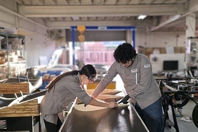 Two people in gray work shirts bend over a long, curved panel on a workbench in a cluttered workshop with shelves and equipment. One is smoothing or fitting the piece while the other holds it in place.