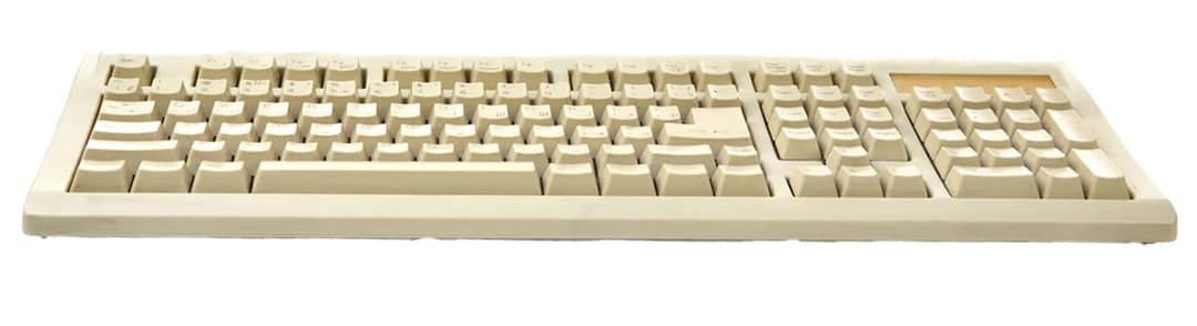 The image shows a beige computer keyboard with a slightly curved design, resting on a black background. The keyboard features a standard QWERTY layout with white keys and black lettering. The keys are arranged in a standard QWERTY pattern, and the keyboard has a slightly raised beige plastic or rubber top. The keyboard