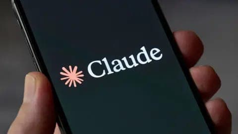 The image shows a hand holding a black smartphone. The screen of the smartphone displays the word "Claude" in white text, with a pink flower-like logo positioned to the left of the text. The background is blurred, drawing focus to the smartphone and the word "Claude".
