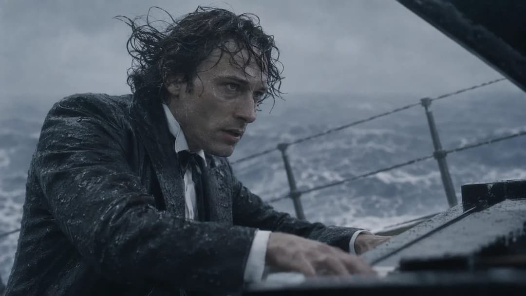 The image shows a man seated at a piano on a ship, with the piano lid open. He is dressed in a dark suit and tie, and his hair is wet and appears to be blowing in the wind. The ship's deck is visible in the background, with a dark, possibly black, railing. The ocean is visible in the background, with waves crashing aga