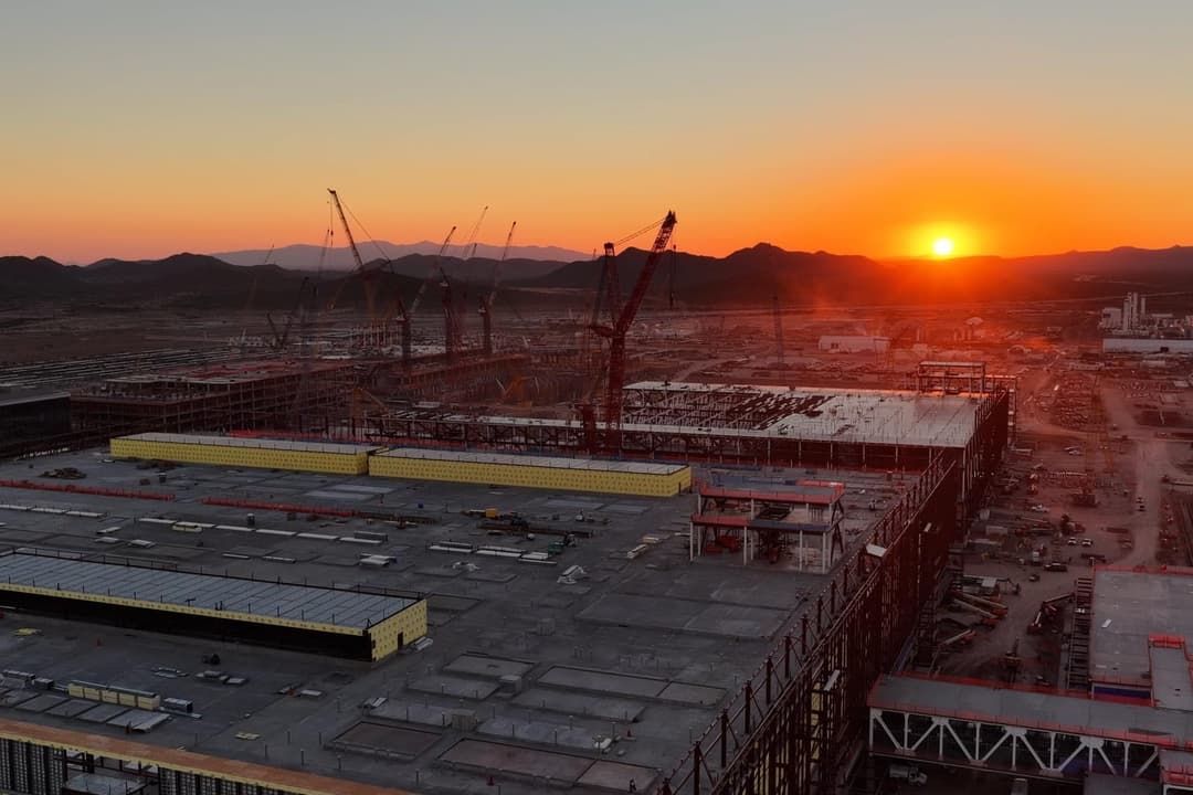Aerial view at sunset of a large construction site with steel-framed buildings, yellow-walled structures, and tall cranes, with desert mountains on the horizon. No legible text.