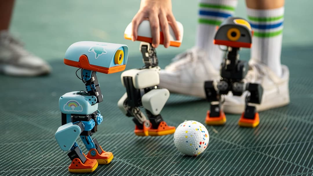 The image shows three small robots on a green surface, possibly a tennis court. The robot on the left is blue and white with a rainbow design on its body and orange accents. The middle robot is primarily white with orange accents and a yellow/orange camera on its head. The robot on the right is primarily black and oran