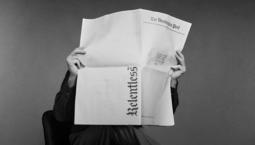 The image shows a person holding a newspaper, which is the Washington Post, in their hands. The person is wearing a dark-colored, possibly black, long-sleeved shirt. The newspaper is open, revealing the headline "The Washington Post" at the top. The person's face is obscured by the newspaper, and the image is in black