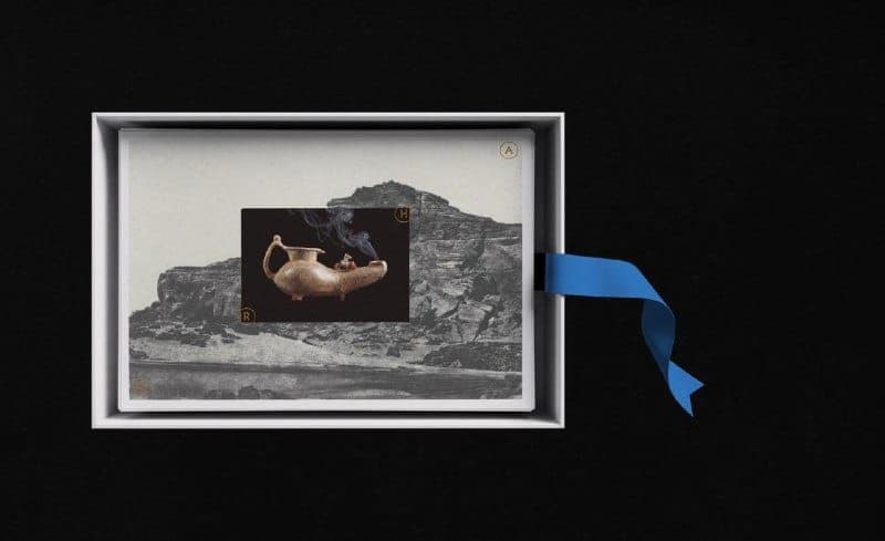 The image shows a white box with a blue ribbon attached, resting on a black surface. The box contains a black and white photograph of a landscape featuring a rocky cliff and a small, possibly golden, vessel emitting smoke. The photograph is framed by a white border. The word "A" is visible in the top right corner of th