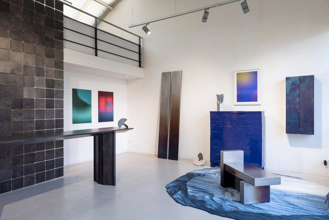 The image depicts a modern art gallery with a high ceiling and a black metal railing overlooking a second-floor balcony. The gallery features a minimalist aesthetic with white walls and a gray concrete floor. A black metal table with a dark surface is positioned in the foreground, and a blue cabinet with a dark surface