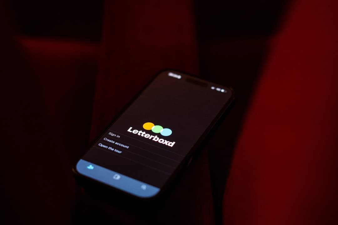 The image shows a black smartphone resting on a dark red fabric surface, possibly a theater seat. The phone's screen is illuminated, displaying the Letterbox app. The app's logo, featuring three colored circles, is prominently displayed on the screen. The screen also shows a menu with options for "Sign in", "Create acc