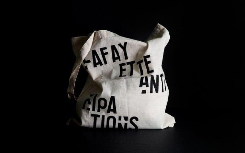 The image shows a white canvas tote bag with a black text design. The bag is slightly crumpled and has a light beige or cream-colored handle. The text on the bag is arranged in a diagonal pattern and reads "FAFAY ETTE ANTI". The bag is positioned against a black background, which highlights its white color and black te