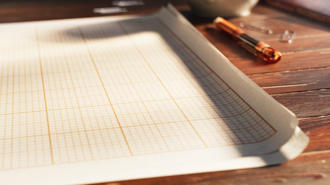 The image shows a wooden desk with a white paper featuring a grid pattern, likely a graph paper, resting on a wooden surface. A pen with a gold-colored body and a dark-colored tip is positioned to the right of the paper. A few small, clear plastic clips are scattered near the pen. The desk is bathed in sunlight, castin