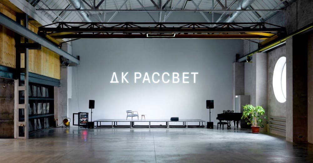 The image depicts a spacious, industrial-style room with a high ceiling and exposed metal beams. The room features a large white wall with the text "��K PACCBET" in white letters. A long, dark-colored stage is positioned in the center of the room, with a few pieces of furniture, including a chair and a small table, pla