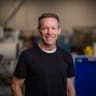 The image shows a man standing in a warehouse setting. He is wearing a black t-shirt and has a friendly smile on his face. The warehouse features a blue wall and a variety of industrial equipment in the background, including a yellow object. The man's face is the central focus of the image, and he is positioned slightl