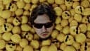 The image shows a man with dark brown hair and a mustache, wearing sunglasses, partially submerged in a large pile of yellow lemons. The lemons are arranged in a way that partially obscures the man's face. The man's face is visible, but his expression is not clearly discernible. The lemons are mostly round and yellow,