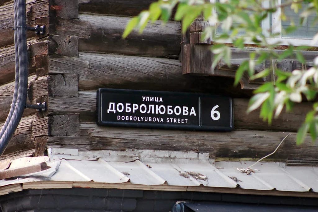 The image shows a black street sign with white text affixed to a wooden building. The sign reads "�������������������� 6 DOBROLYUBOVA STREET". The building features a log wall and a corrugated metal roof, with a window visible on the right side. A tree branch with green leaves is partially visible in the foreground. Th