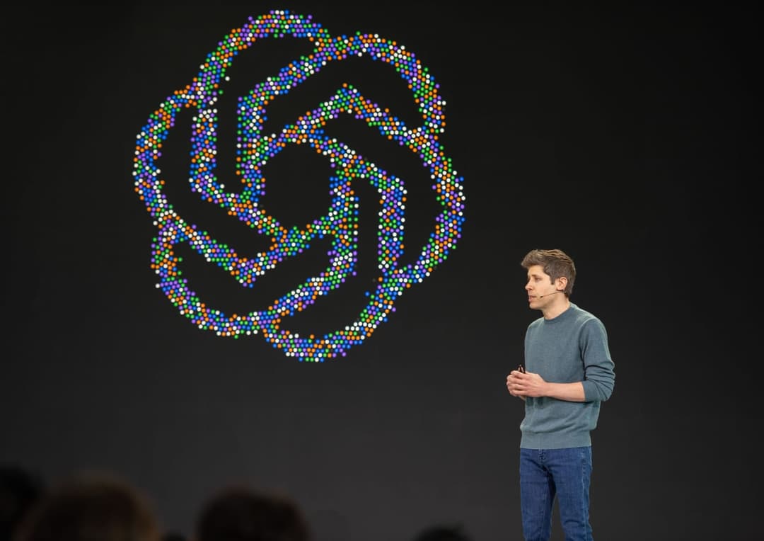 The image shows a man standing on a stage, dressed in a gray sweater and blue jeans. He is gesturing with his hands, possibly explaining something. Behind him, a large, colorful, abstract design composed of small, multicolored dots is displayed on a black background. The design is a complex, swirling pattern. The man's