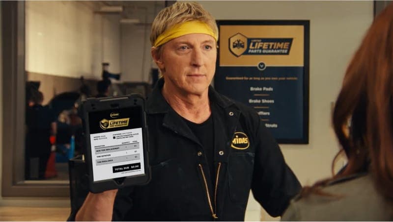 The image shows a man wearing a black jacket with a yellow logo, standing in what appears to be a garage or service area. He is holding a tablet in his left hand and is wearing a yellow headband. The man is wearing a black shirt underneath his jacket. Behind him, a sign for "Lifetime Parts Guarantee" is visible. The si