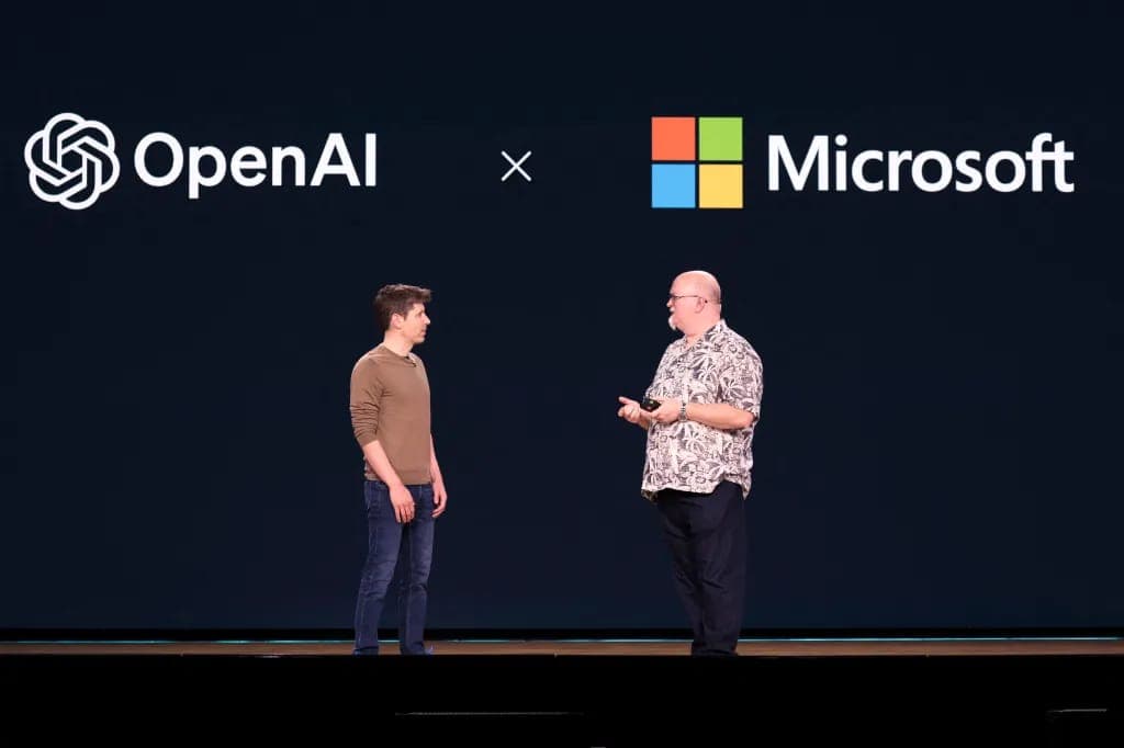 Two men stand on a dark stage in front of a screen showing the OpenAI logo, an "×" sign, and the Microsoft logo. The man on the left wears a brown sweater and jeans, and the man on the right wears a patterned short-sleeved shirt and dark trousers and holds a small device.