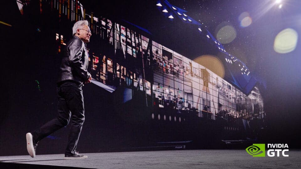 The image shows a man standing on a stage, dressed in a black leather jacket and black pants. He is holding a microphone and appears to be speaking or presenting. The stage is illuminated by a large screen displaying a cityscape or a similar scene. The screen is surrounded by a dark background, and the stage floor is a